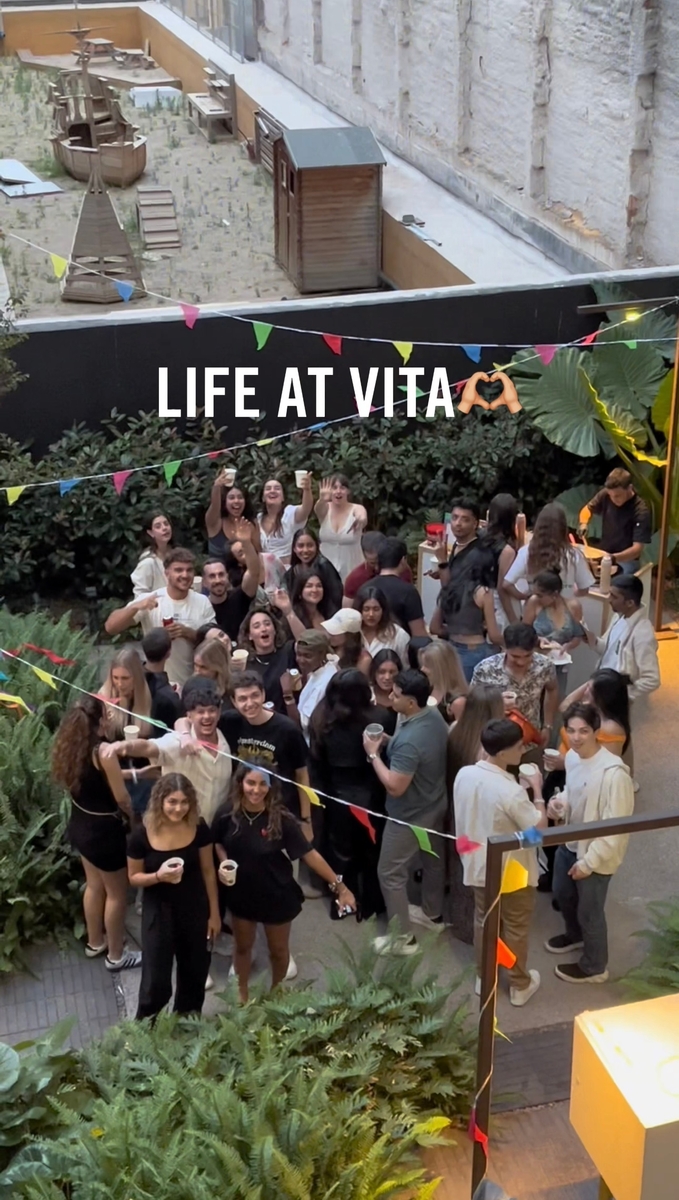 A group of people gathered outdoors, smiling and holding drinks. Colorful bunting hangs above, and lush green plants surround them. A sign reads "LIFE AT VITA" with two raised hands emoji. A courtyard with a sandbox and structure is visible in the background.